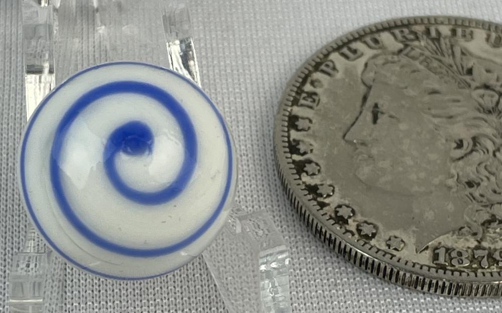 Close-up of a white marble with blue swirl rings and a clear blue dot center, shown next to a silver coin on a textured surface.