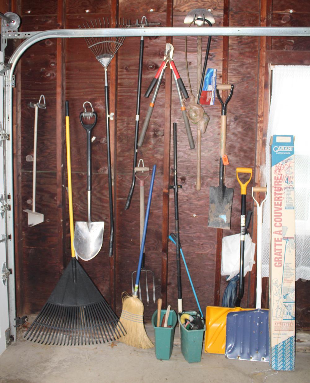 Assortment of garden hand tools hanging on hooks in a storage shed, including shovels, rakes, and brooms.