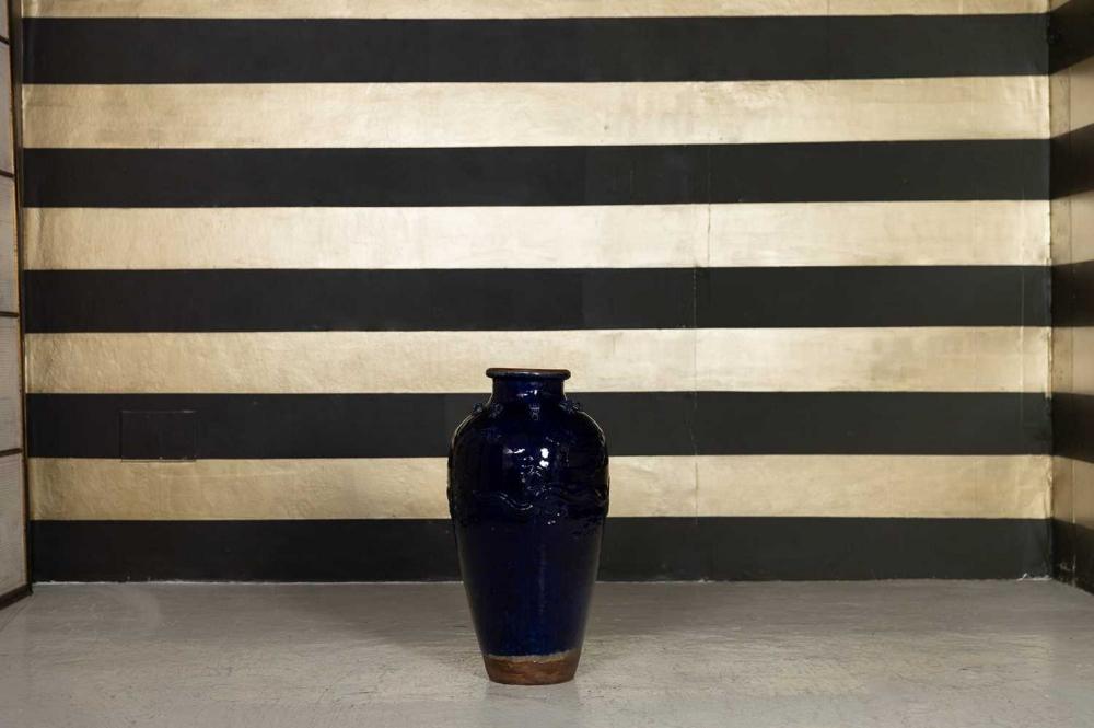 Blue-glazed ceramic jar with a narrow neck and flared rim, photographed against a striped wall.