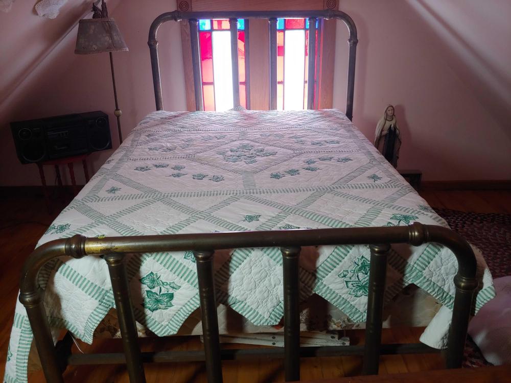 Full-size bed with a brass-colored metal headboard and footboard, covered by a quilt with green and white pattern.