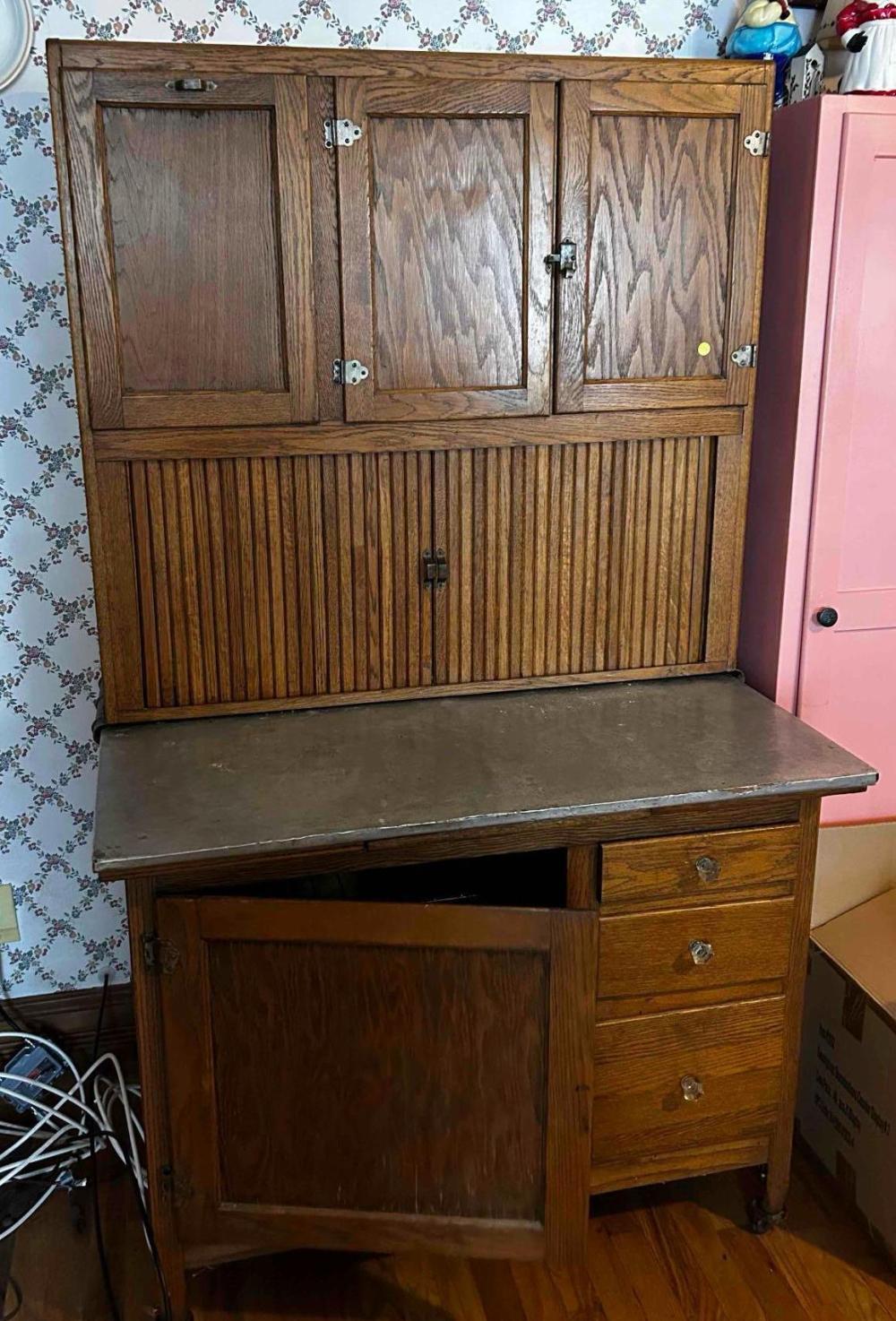 Wooden hoosier cabinet with hinged doors and a metal countertop, shown against a floral wallpaper background.