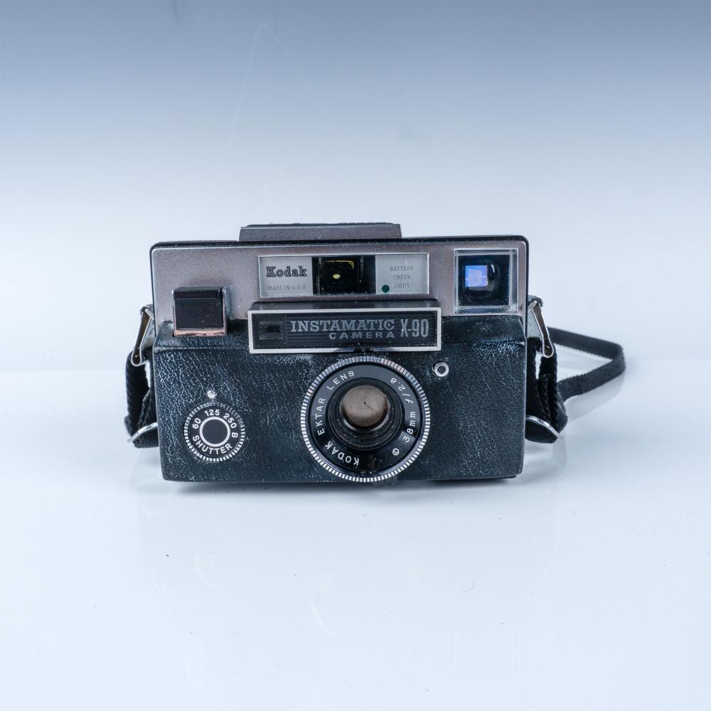 Front view of a Kodak Instamatic X-90 camera with a black strap, showing the lens and shutter speed dial.