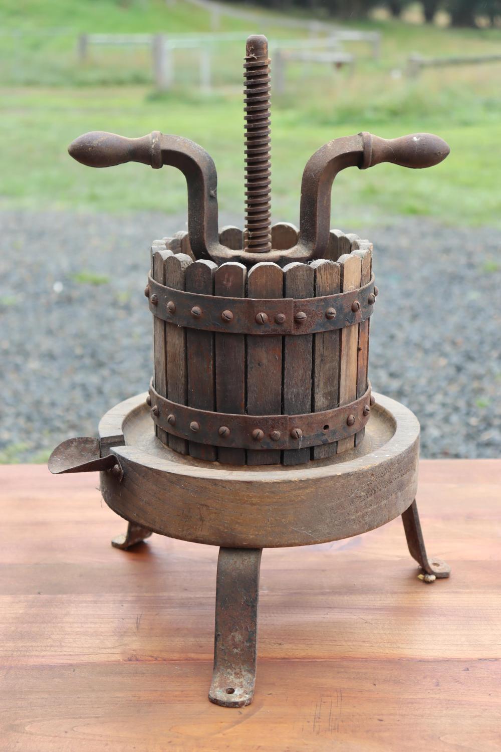 Antique cider press with iron screw mechanism and wooden barrel container on three metal legs, outdoors background.