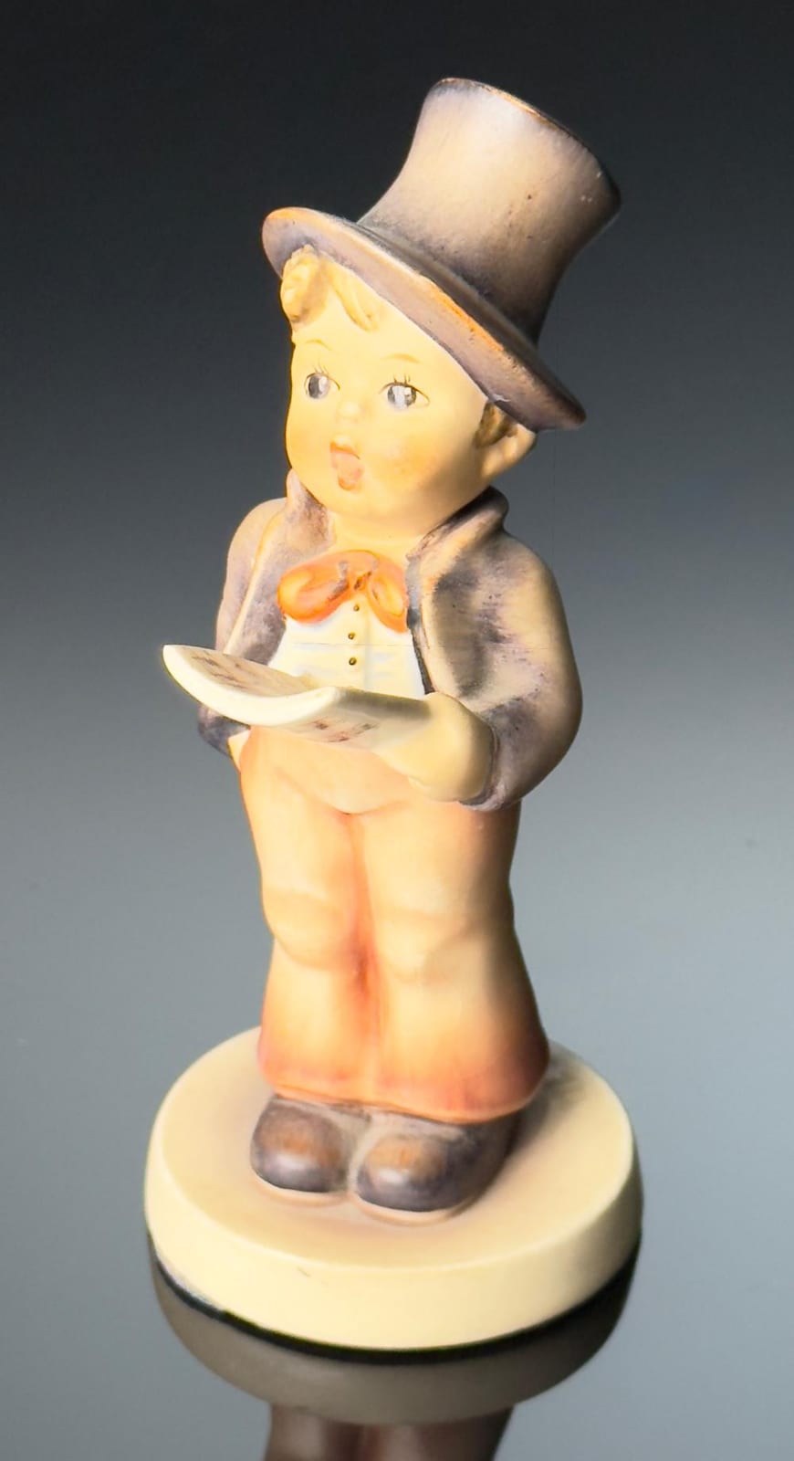 Small painted figurine of a child holding an orange musical instrument, wearing a white shirt and a tall dark hat, on a round base.