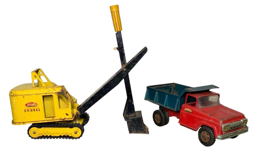 Two small toy construction vehicles: a yellow tracked shovel and a red dump truck with a teal trailer, shown with a black metal shovel attachment in the center.