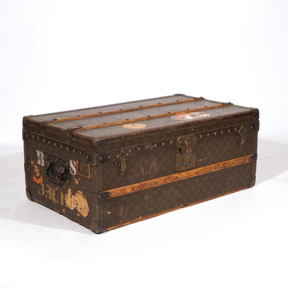 Brown vintage steamer trunk with riveted metal trim, leather straps, and decorative stickers on the front and sides.