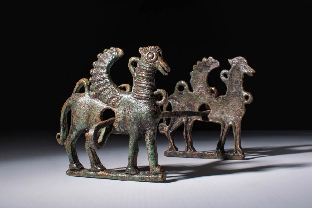 Bronze horse bit with two standing horses and winged horse motifs, shown on a dark background.