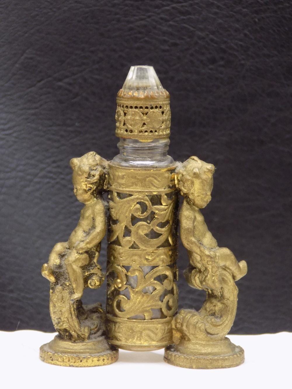 Ornate small perfume bottle with a clear cap and gold-colored decorative cupid figures on both sides.