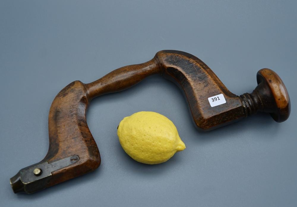 Antique-style wooden brace hand drill with metal bits and a yellow lemon placed on a blue background.