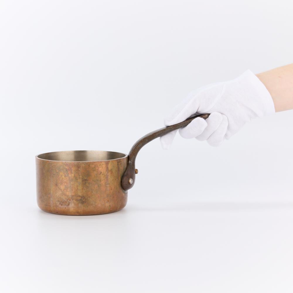 Hand holding the curved handle of a small copper sauce pot with side rivets.