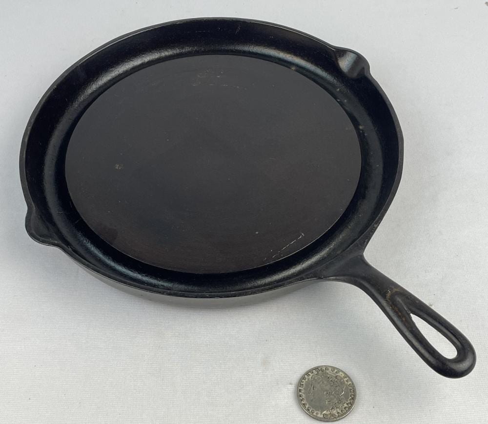 Black cast iron frying skillet with loop handle, shown on a light background with a small coin for size comparison.