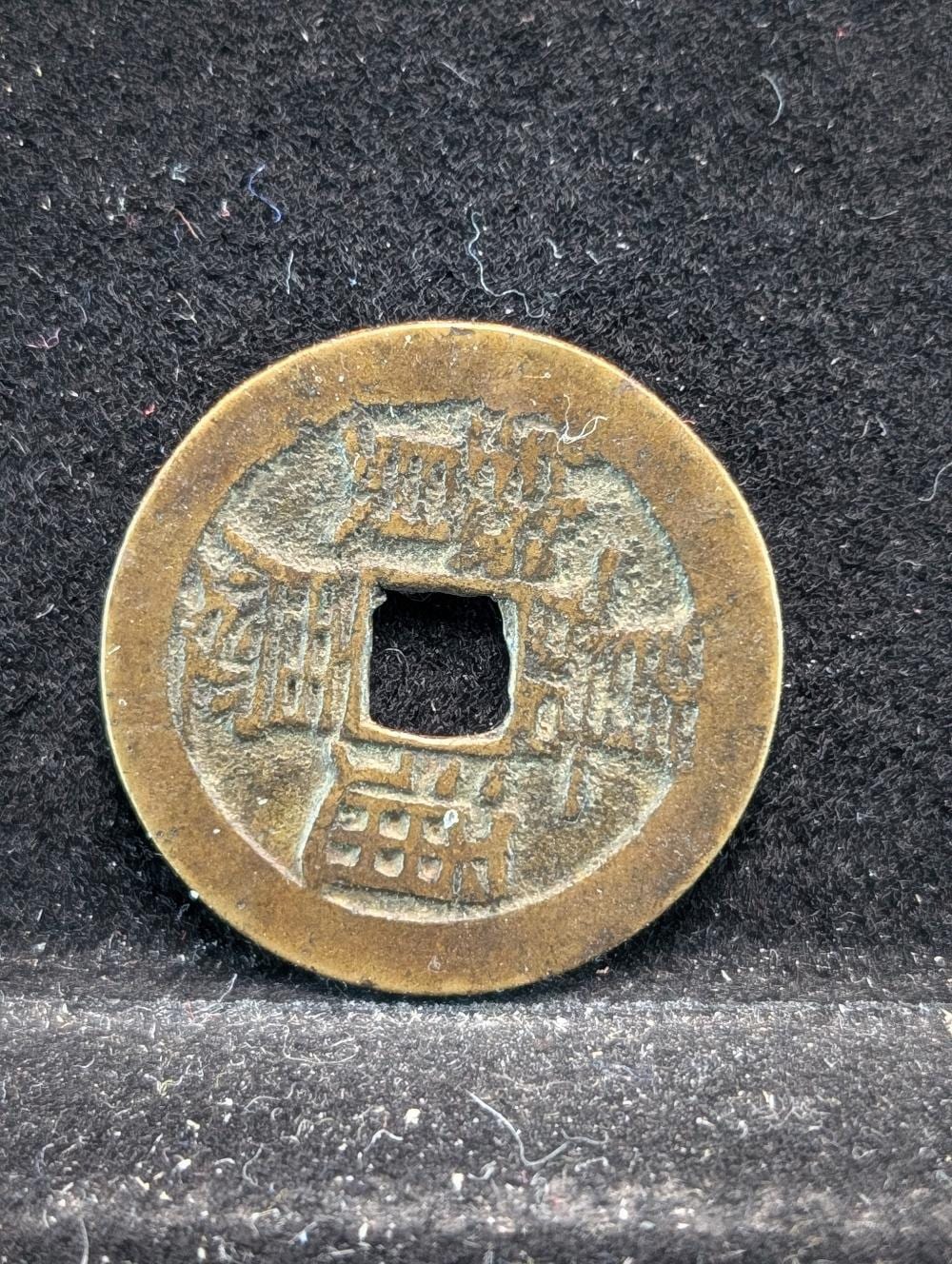 Close-up of a round gold-colored Chinese cash coin with square center hole, shown on a dark textured surface.