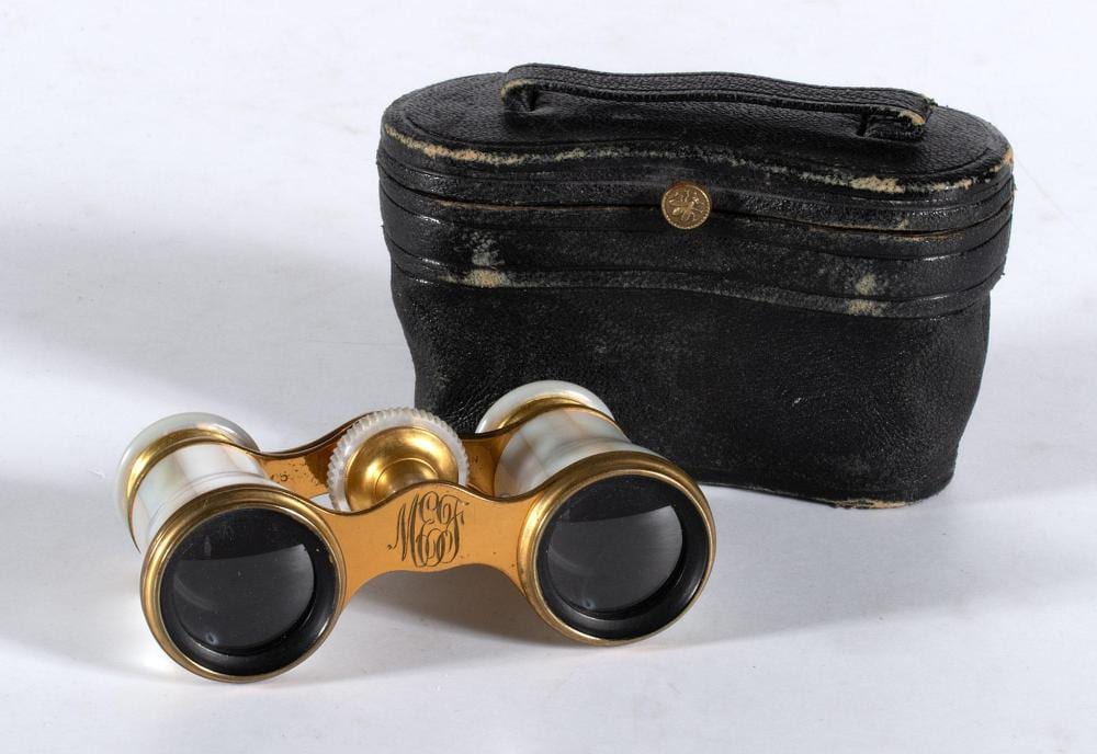 Pair of gold-tone opera glasses with white decorative eyecups resting beside a black leather case.