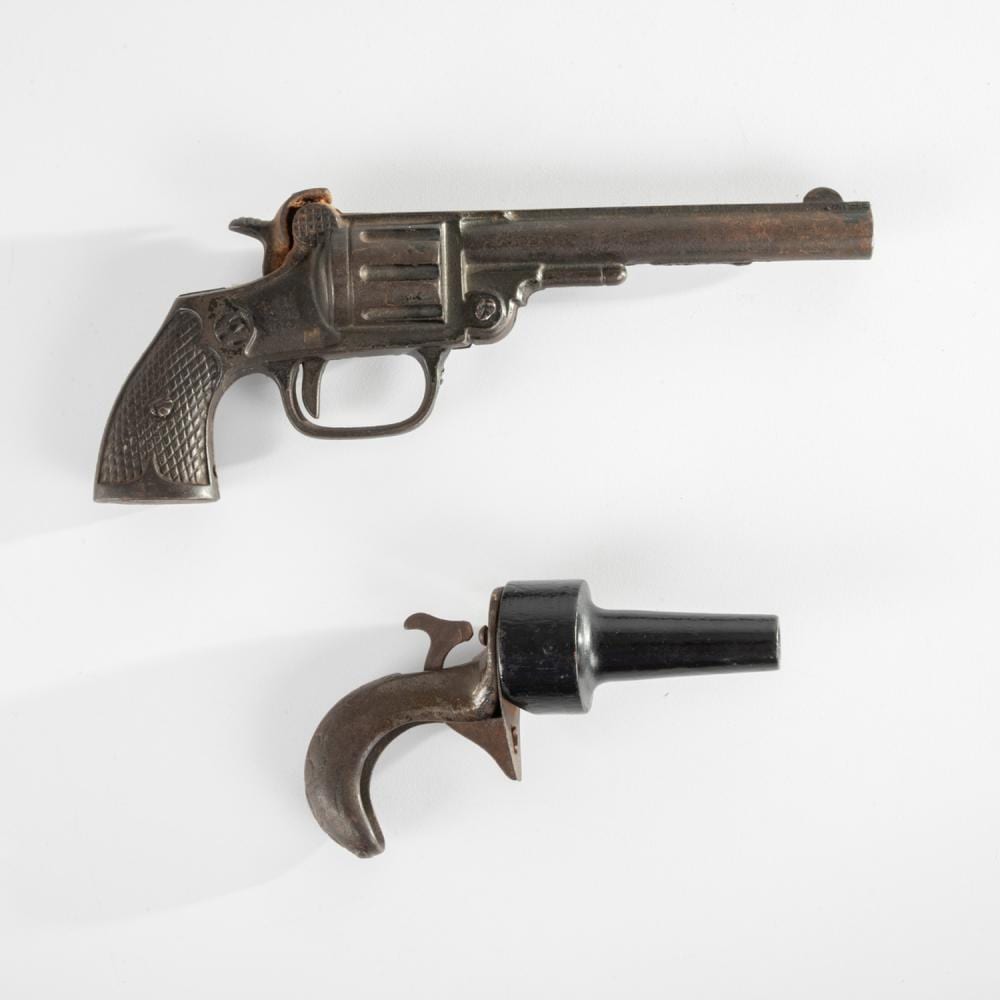 Two cast-iron toy guns with dark brown and black finishes shown on a white background, one lying horizontally and one upright with a curved trigger guard.