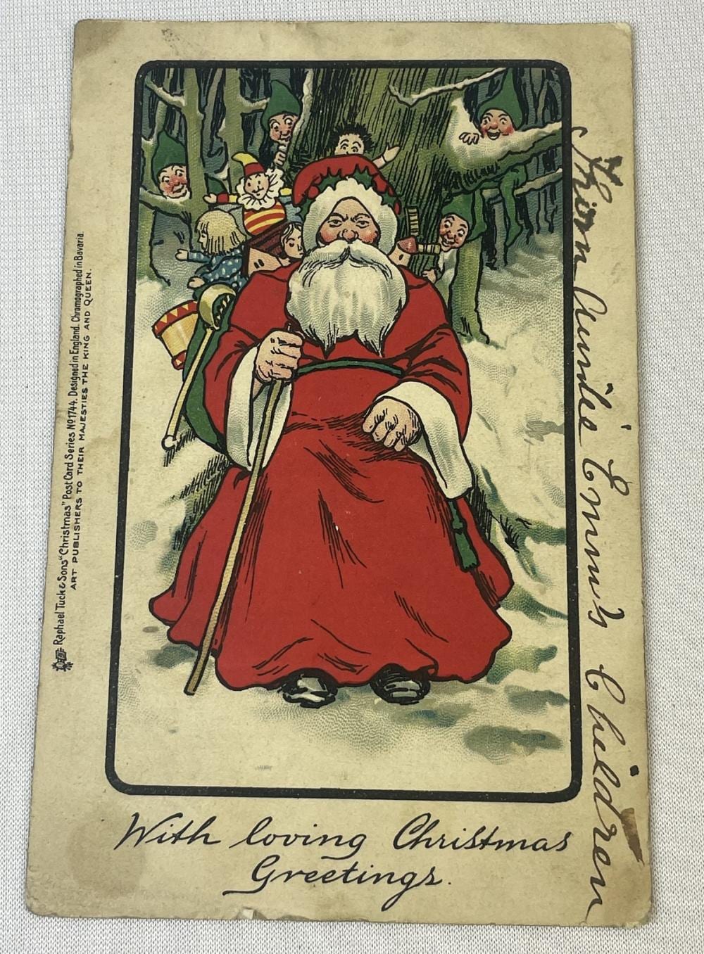 Color illustration on a vintage postcard of Santa Claus sitting in a forest with elves in the trees, holding a letter; cursive text below.