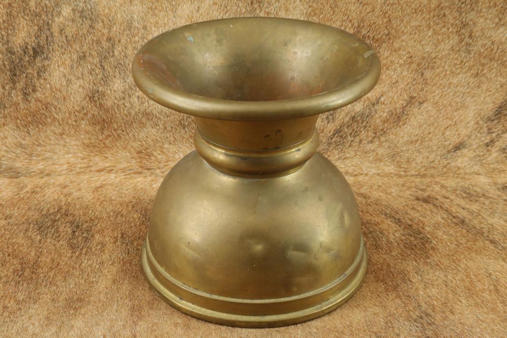 Brass-colored metal spittoon with a flared rim and rounded base shown on a textured surface.