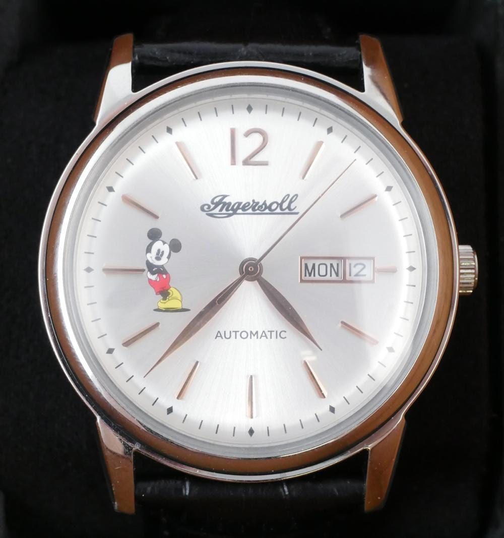 Close-up of a round wristwatch face with the brand “Ingersoll,” Mickey Mouse graphic, and “AUTOMATIC” text; hands, hour markers, and date display are visible.
