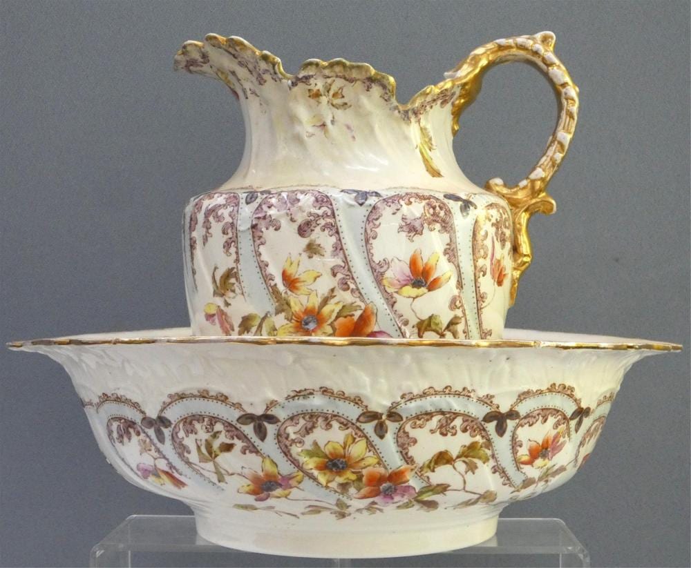 Porcelain wash basin and pitcher set with floral panels and gilded gold handle, photographed against a gray background.
