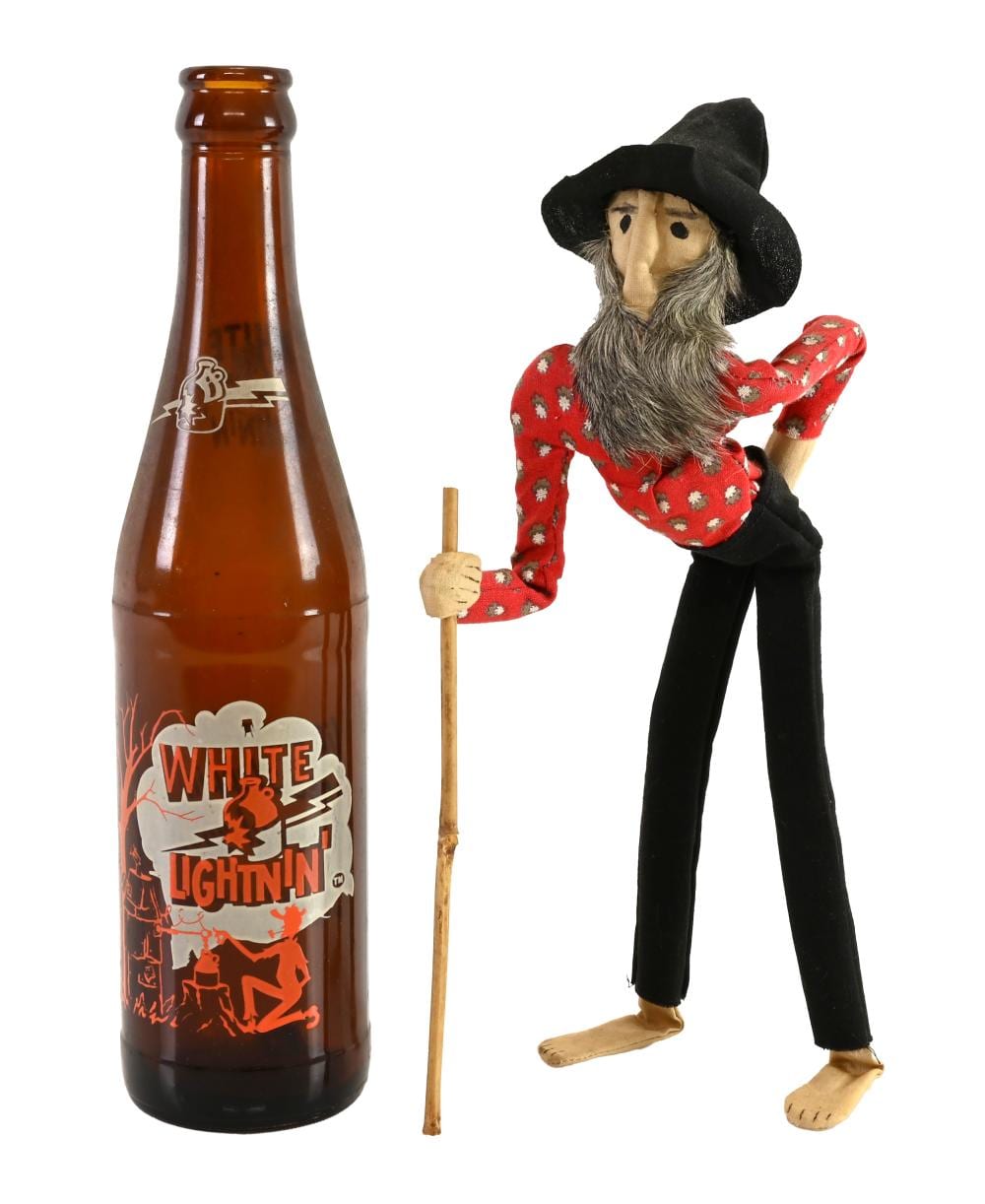 Amber soda bottle labeled “White Lightnin’” next to a hillbilly-style doll with a black hat, gray beard, red patterned shirt, and black pants holding a stick.