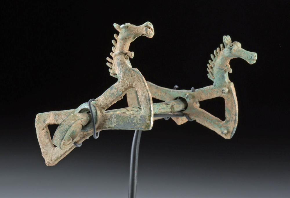 Patinated bronze horse bit with horse-head terminals and ring attachment on a dark background.