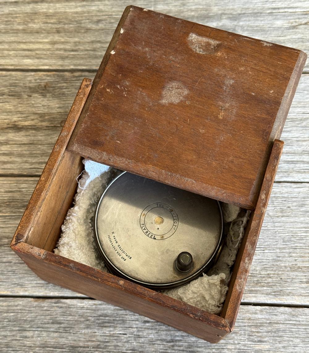 Wooden box open to reveal a metal fishing reel with a circular center plate labeled “The Perfect Fly Reel.”