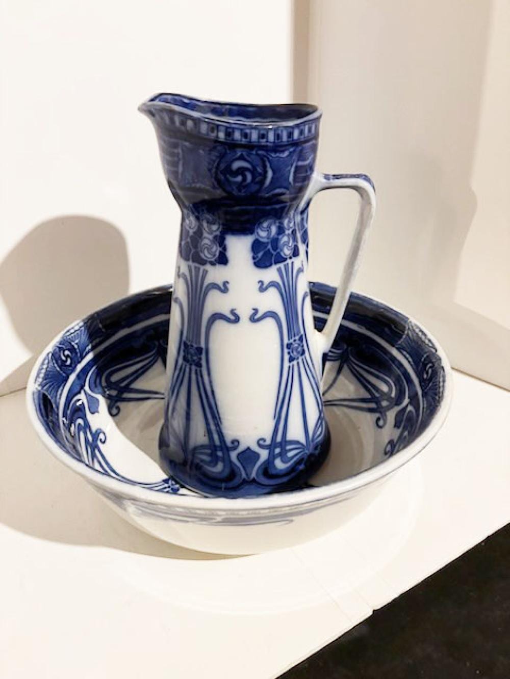 Blue-and-white ceramic pitcher with floral scroll pattern and flowing blue glaze, shown standing on a matching basin.