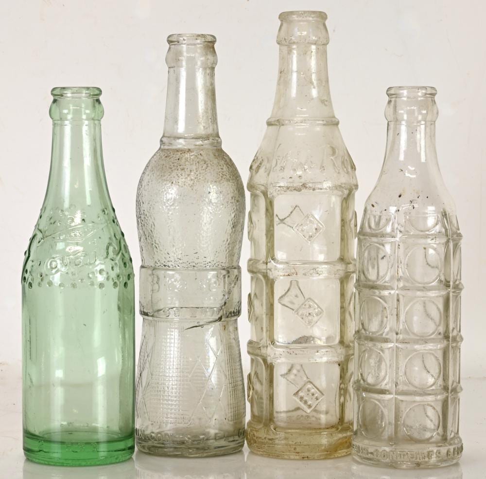 Four vintage glass soda pop bottles in green, clear, and amber colors arranged side by side on a light background.