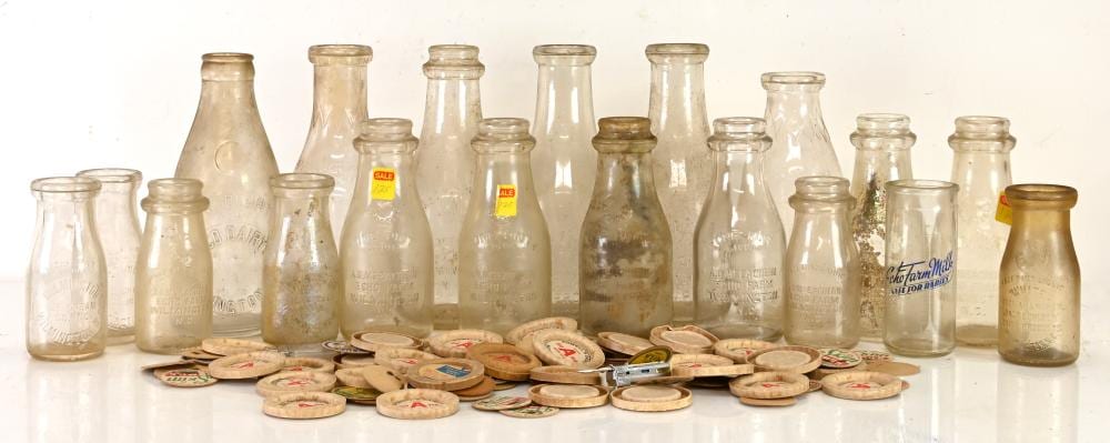 A group of vintage milk bottles of different shapes and colors with a tray of round metal or cardboard bottle caps in front.