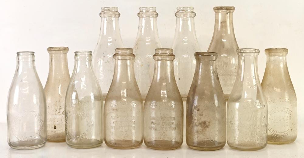 Collection of vintage glass milk bottles in different colors and heights, photographed side by side against a light background.