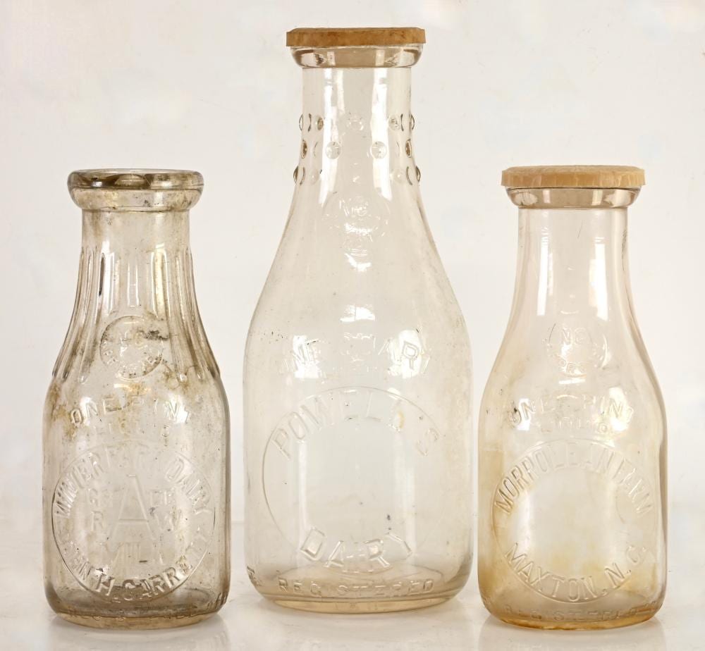 Three vintage clear glass milk bottles with amber screw tops and embossed designs on the front.