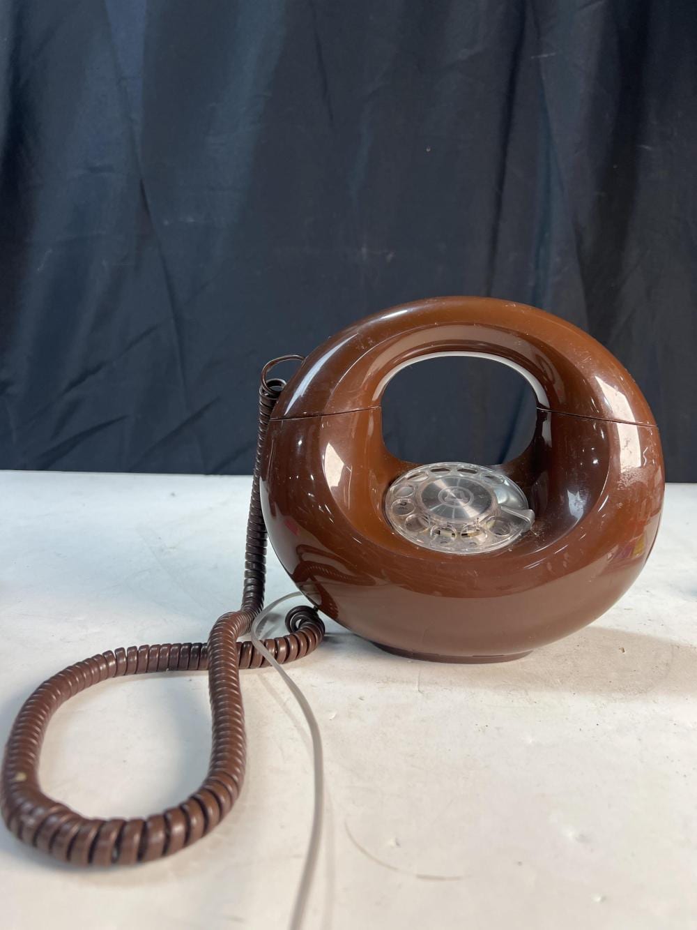 Brown rotary dial “donut” desk telephone with clear dial window and coiled handset cord, photographed against a dark blue backdrop.