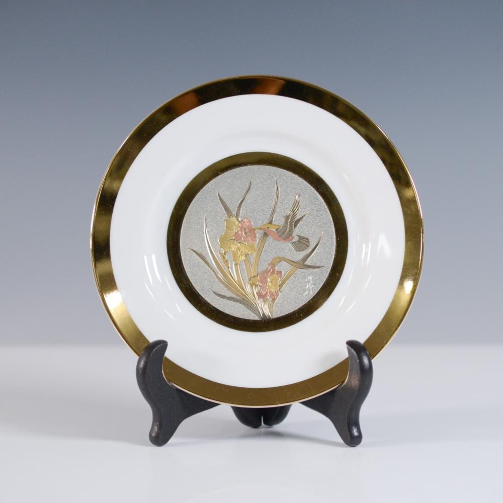 Decorative round plate with white center, gold rim, and a central bird-and-flowers motif in raised metallic decoration, on a stand.