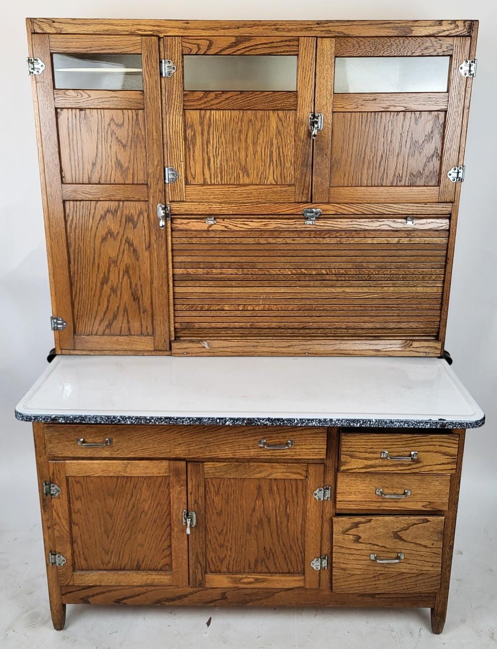 Wood hoosier-style cabinet with glass-front upper doors and multiple drawers, shown with top closed and hinges and handles visible.