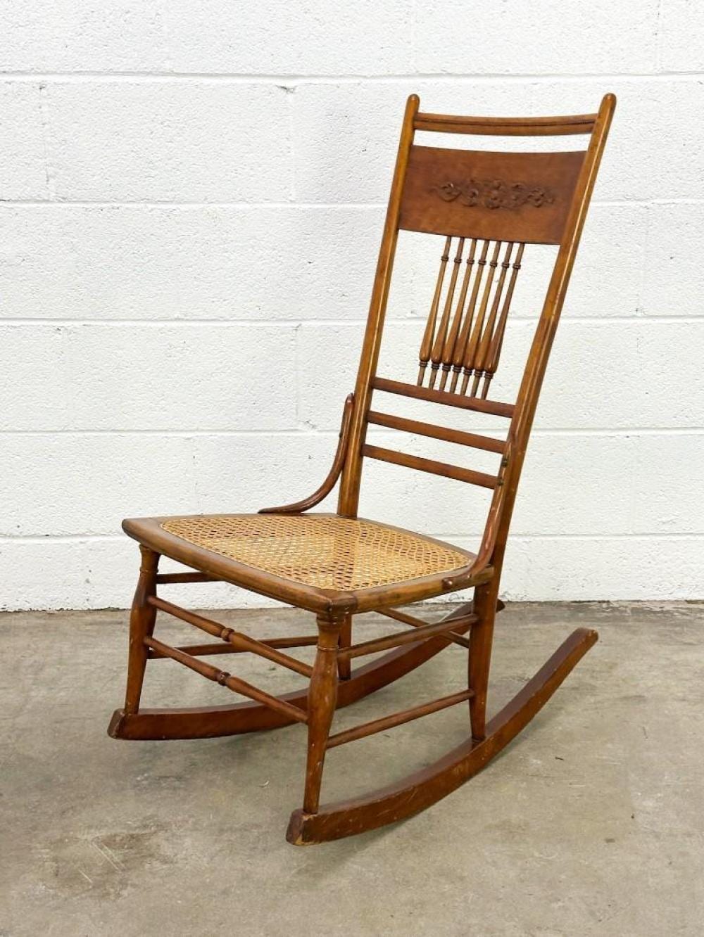 Antique-style wooden rocking chair with a tan woven seat and a carved back panel, shown against a white wall.