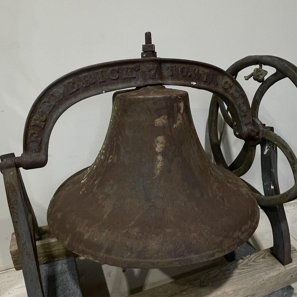 Close-up of a large dark cast iron dinner bell with a flared bell body, wheel-and-frame handle, and stamped markings along the rim.