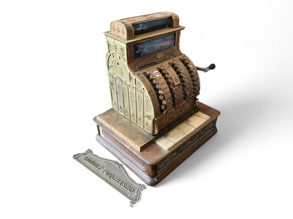 Antique metal cash register with ornate front panel and a drawer, shown with a mounted “Money Purchased” tag plate in front.