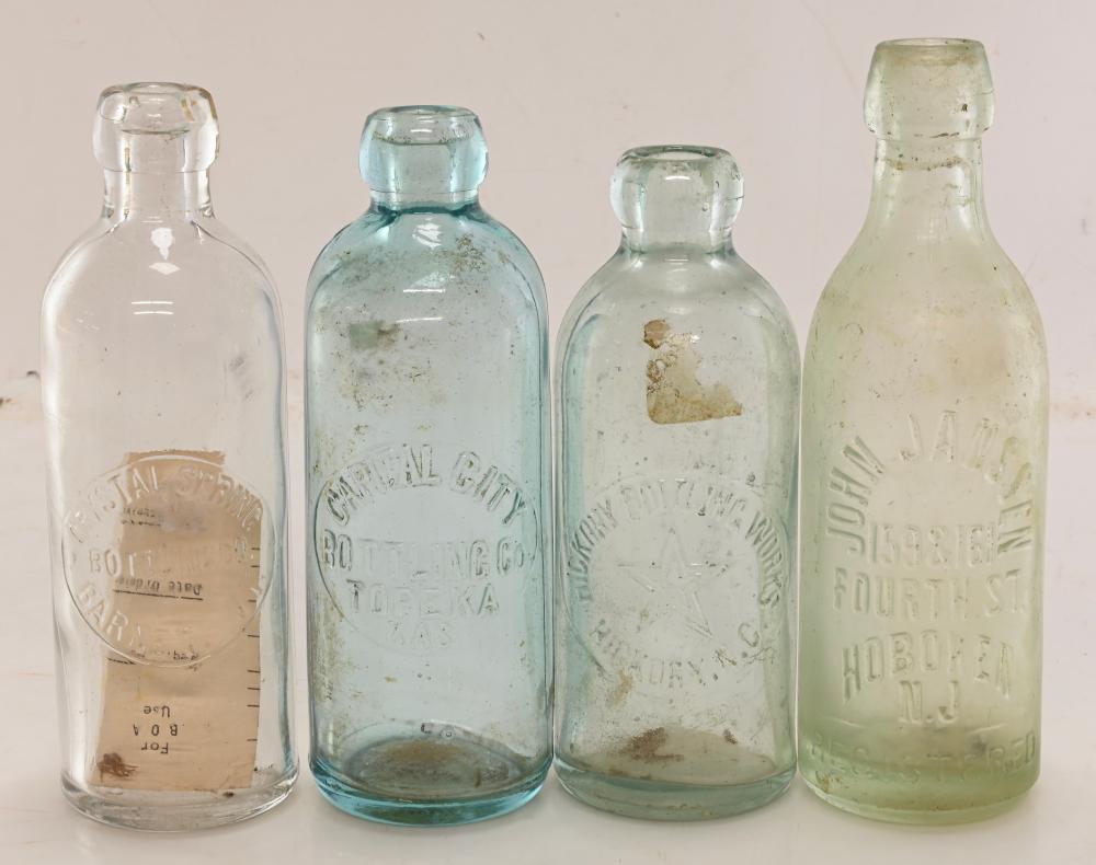 Four antique glass bottles with embossed labels, one clear, one aqua-blue, and two pale green tones, photographed against a white background.