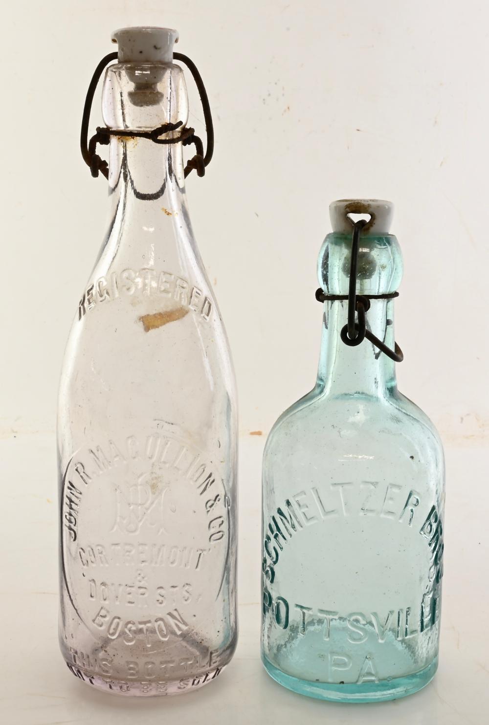 Two antique glass bottles with molded lettering and dark metal carrying handles, one clear and one blue-green.