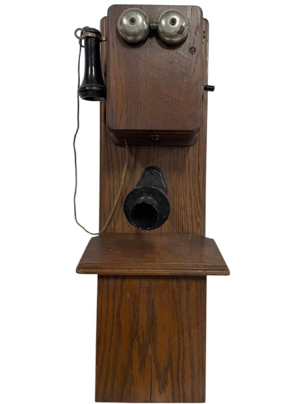 Antique wall telephone with dark wooden cabinet, two silver earpiece covers at the top, and a black handset cradle at center front.