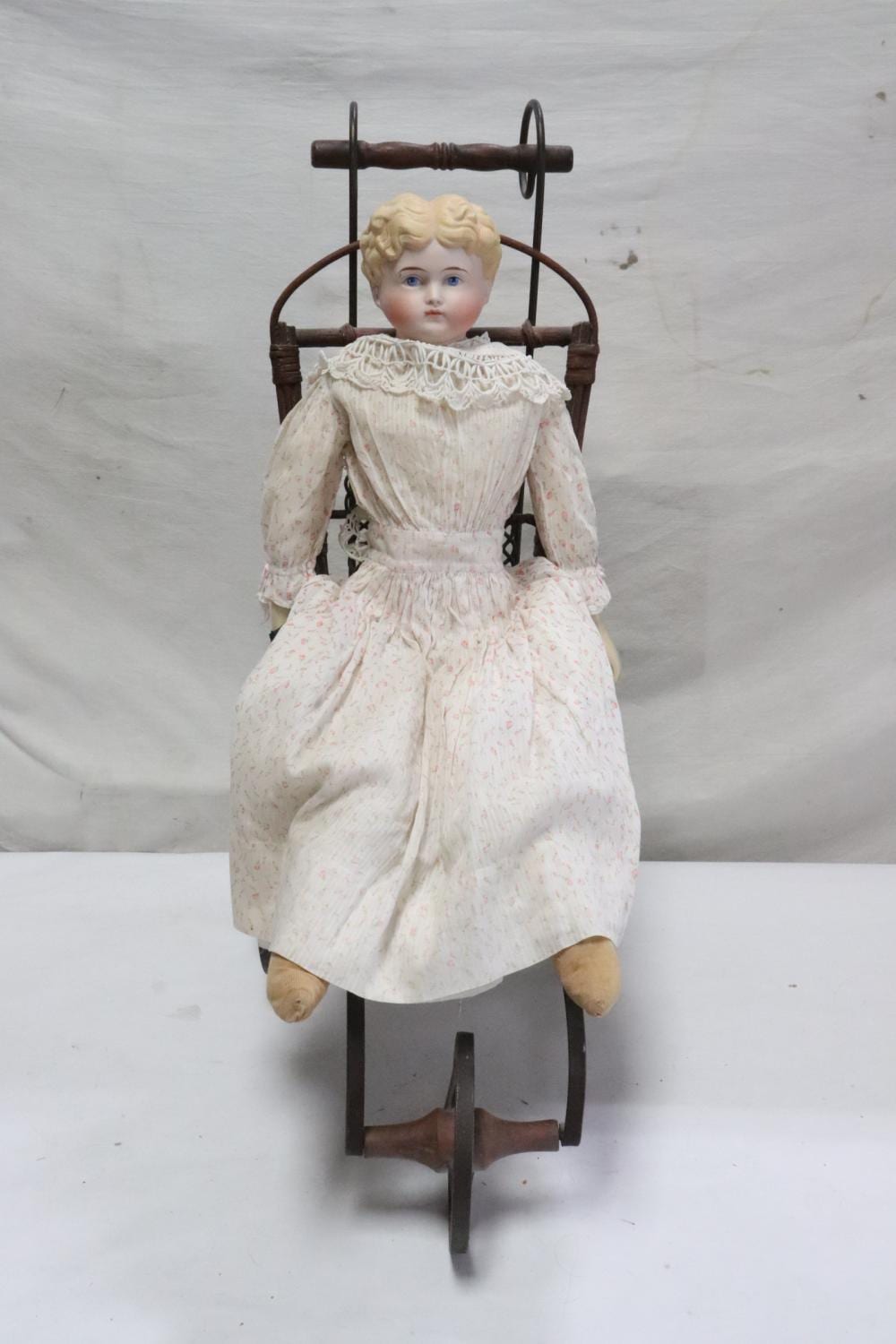 Antique doll seated in a metal doll buggy, with a pale face, curly light hair, and a white lace-trimmed dress.
