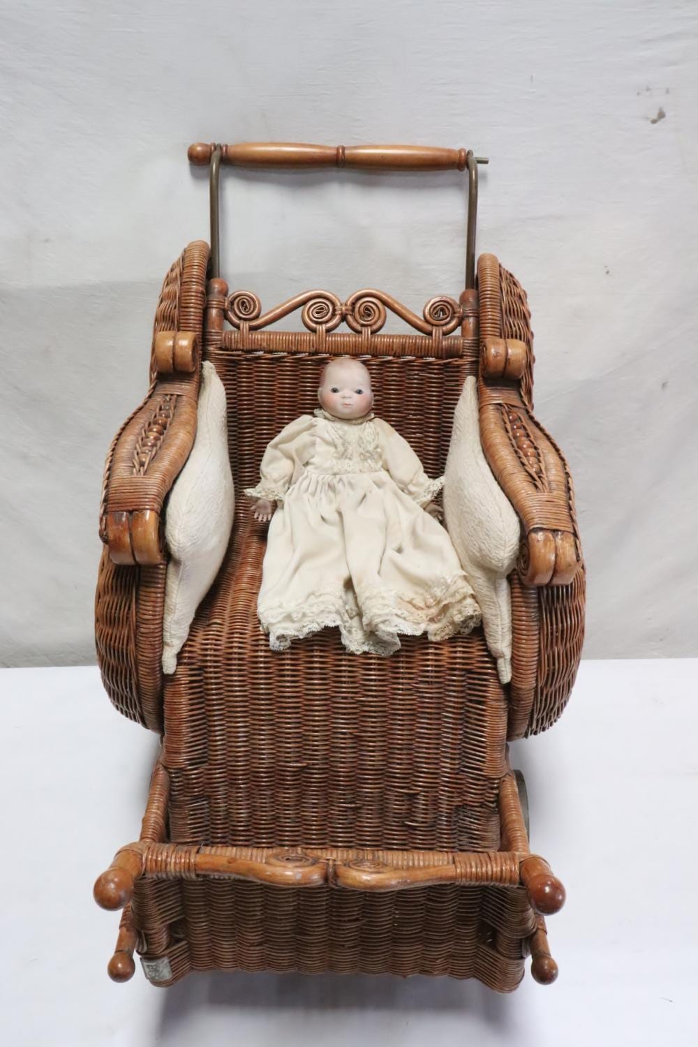 Brown wicker doll buggy with rounded handles and a small baby doll seated in the basket wearing a cream dress and bonnet.