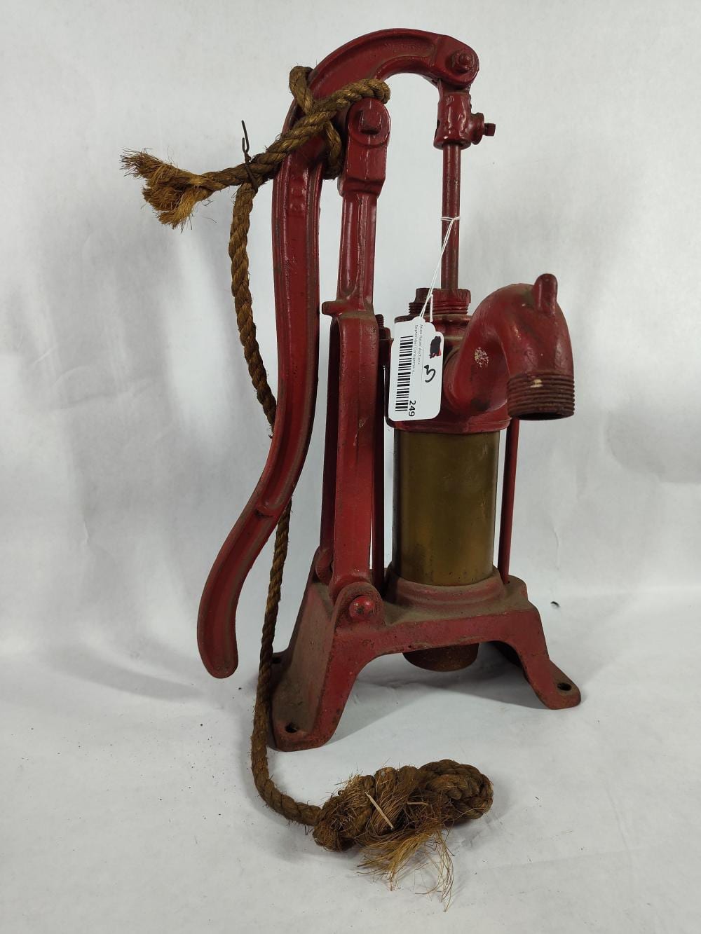 Red and brass hand water well pump with metal lever and rope, photographed on a white background.