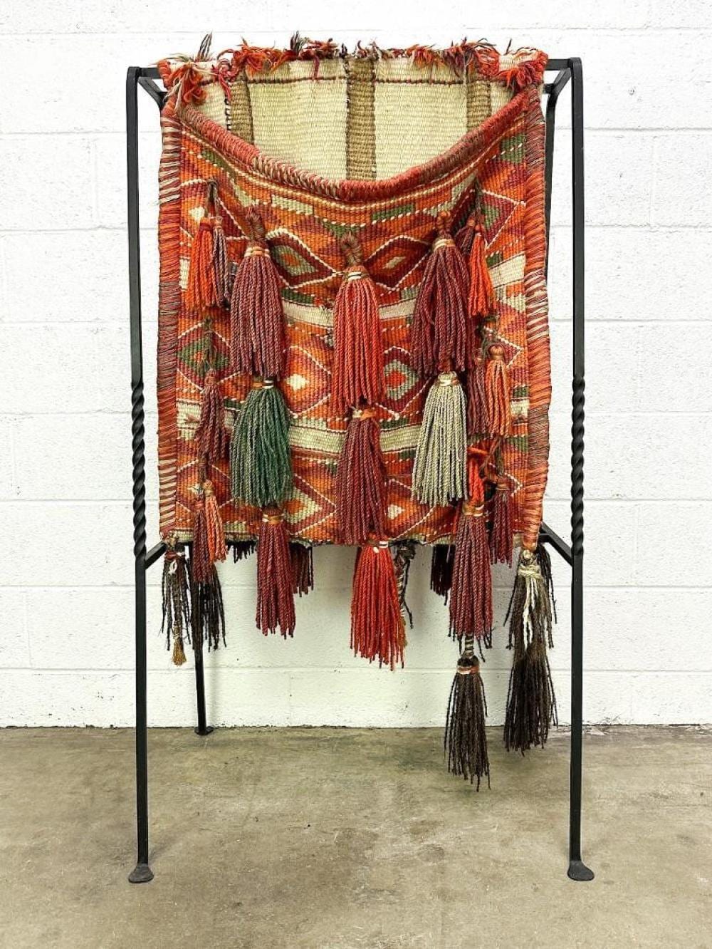 Decorative textile camel saddle cover with tassels and geometric patterns draped on a black wrought-iron stand.