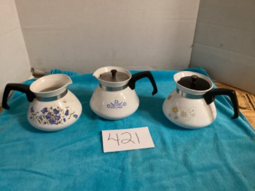 Three white stovetop percolator coffee pots with blue floral patterns and black handles on a teal cloth.