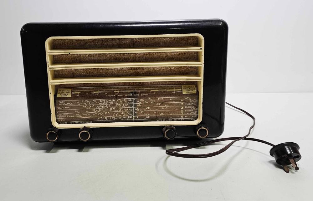 Vintage tabletop radio with black casing, open front speaker grille, station frequency display, and a power cord with plug.
