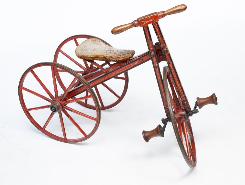 Red and brown painted wooden child’s velocipede tricycle with spoked wheels, seat, and handlebars on a white background.