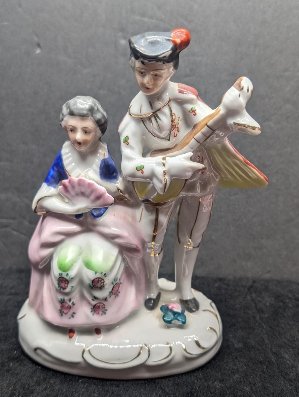Porcelain figurine of a seated woman in a pink dress holding a small object, paired with a standing man in white and gold with a red hat, both on a base with ac