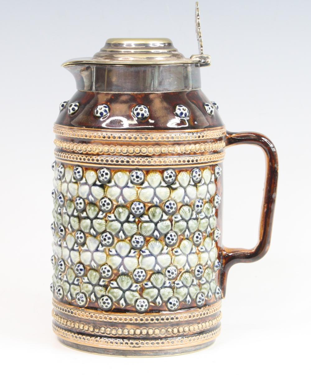 Brown glazed stoneware jug with a lid and handle, featuring raised dot-and-leaf relief decoration in blue and green with a metal collar.