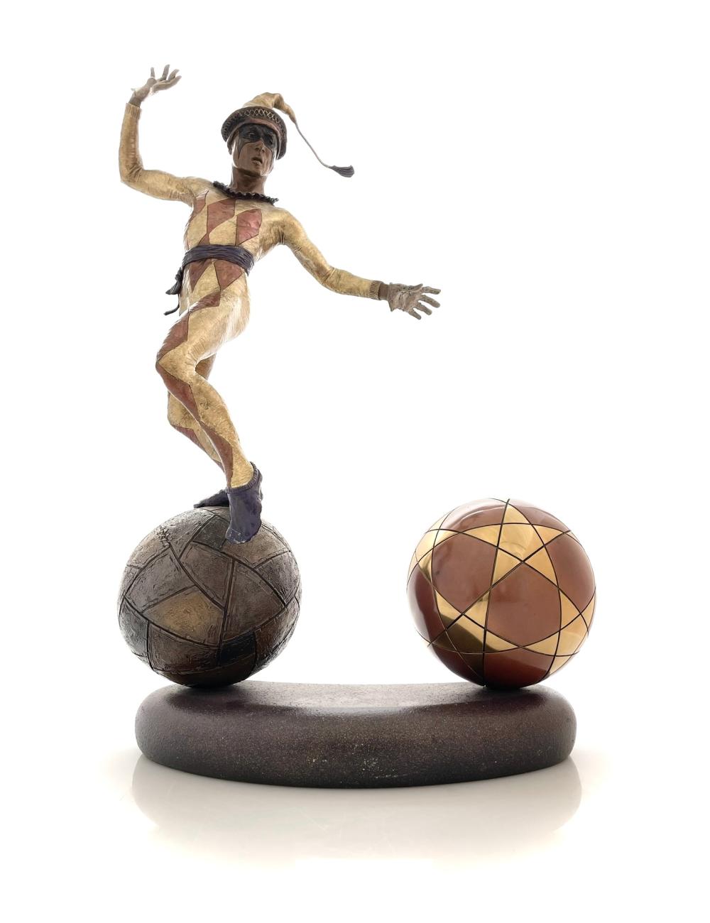 Bronze-like sculpture of a stylized human figure in multicolored patchwork standing on a dark soccer ball and balancing another patterned ball on the base.