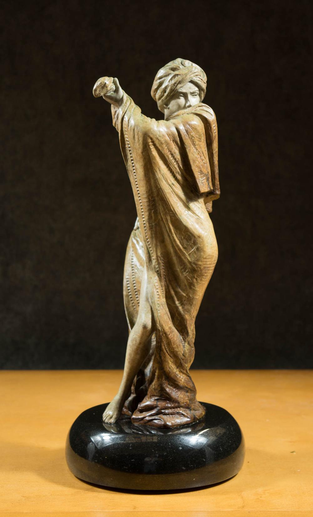 Bronze figurative sculpture of a standing woman with headscarf, raising one arm and pointing forward, on a dark round base.